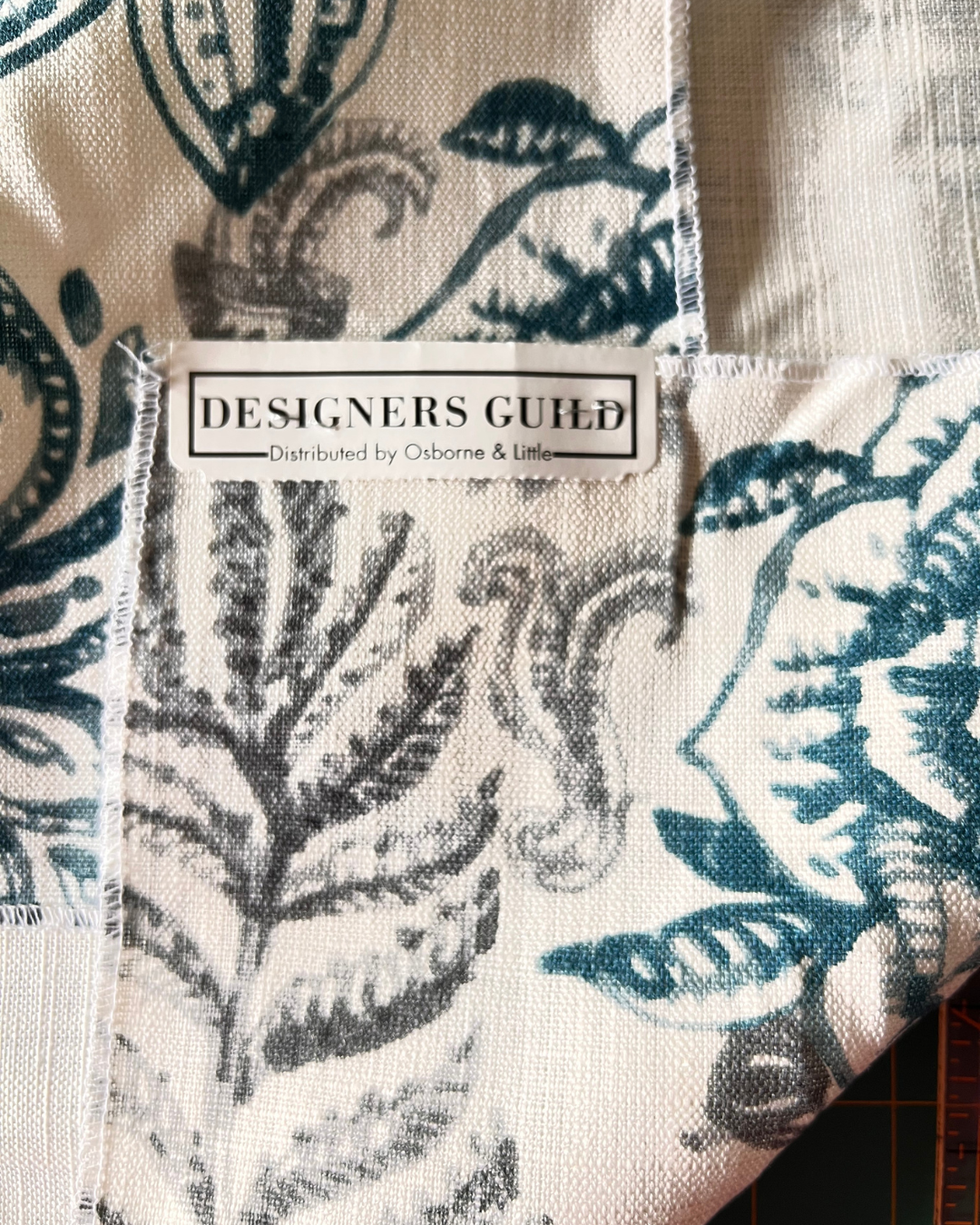 A close-up of a fabric product showing a white woven label that reads 'DESIGNERS GUILD' with 'Distributed by Osborne & Latt', over a botanical print in dark teal and gray on an off-white background.