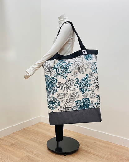 A large tote bag with a blue and gray floral pattern on a white background, displayed on a mannequin in a minimalist room with white walls and light wood flooring.