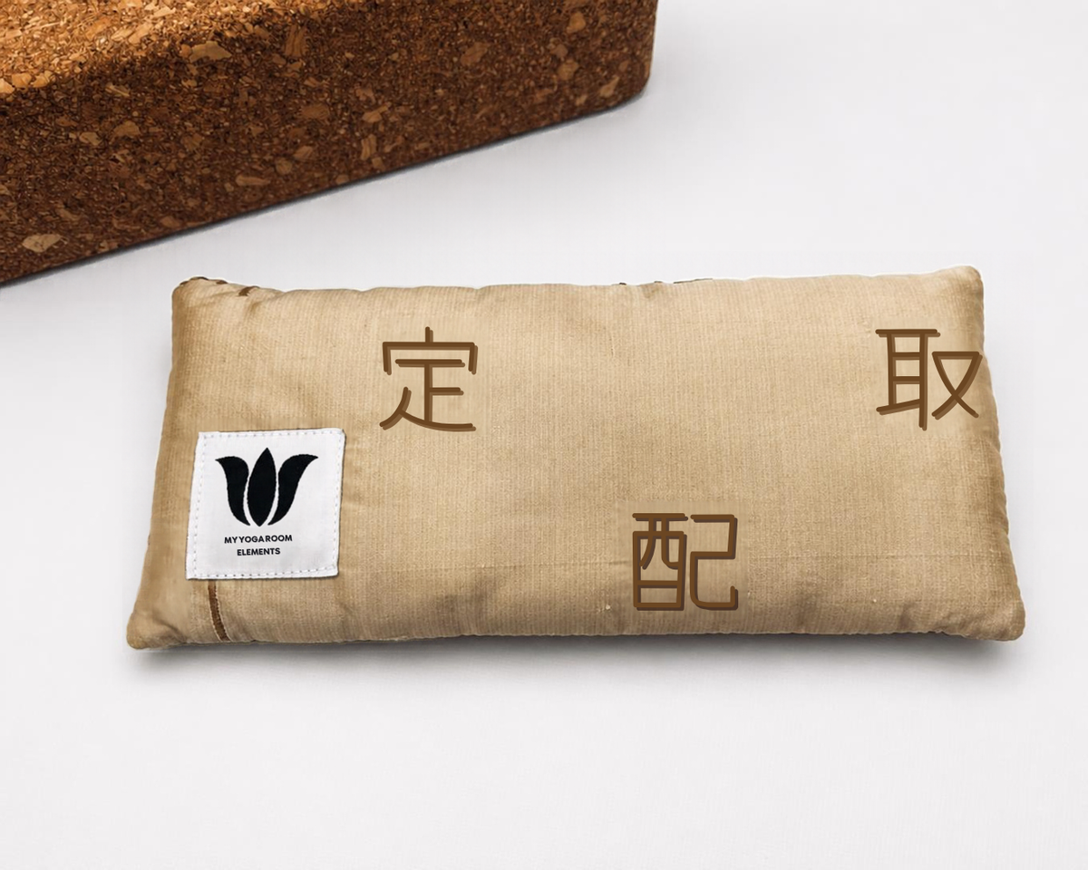 A gold fabric eye pillow with gold asian characters and a white brand label reading 'MY Yoga Room Elements', shown on a white background with a cork block prop in the top left.