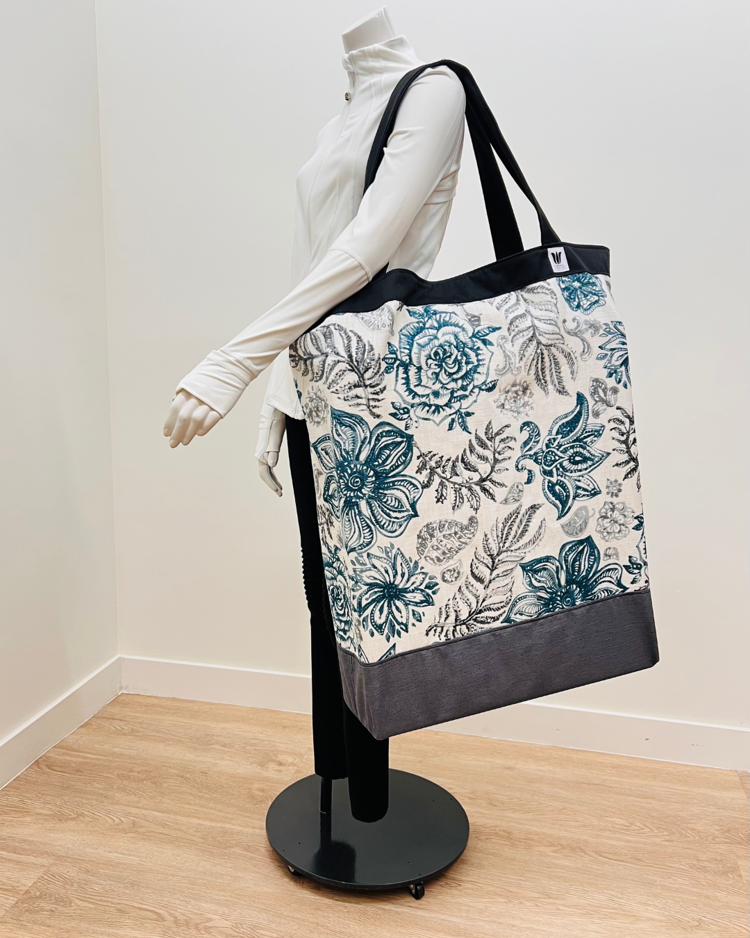 A large tote bag with a blue and gray floral pattern on a white background, displayed on a black mannequin stand in a room with light wood floors and white walls.