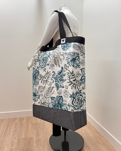 A large tote bag with a blue and gray floral toile pattern on a cream background, featuring black shoulder straps and a dark gray bottom panel, displayed on a mannequin in an indoor setting.