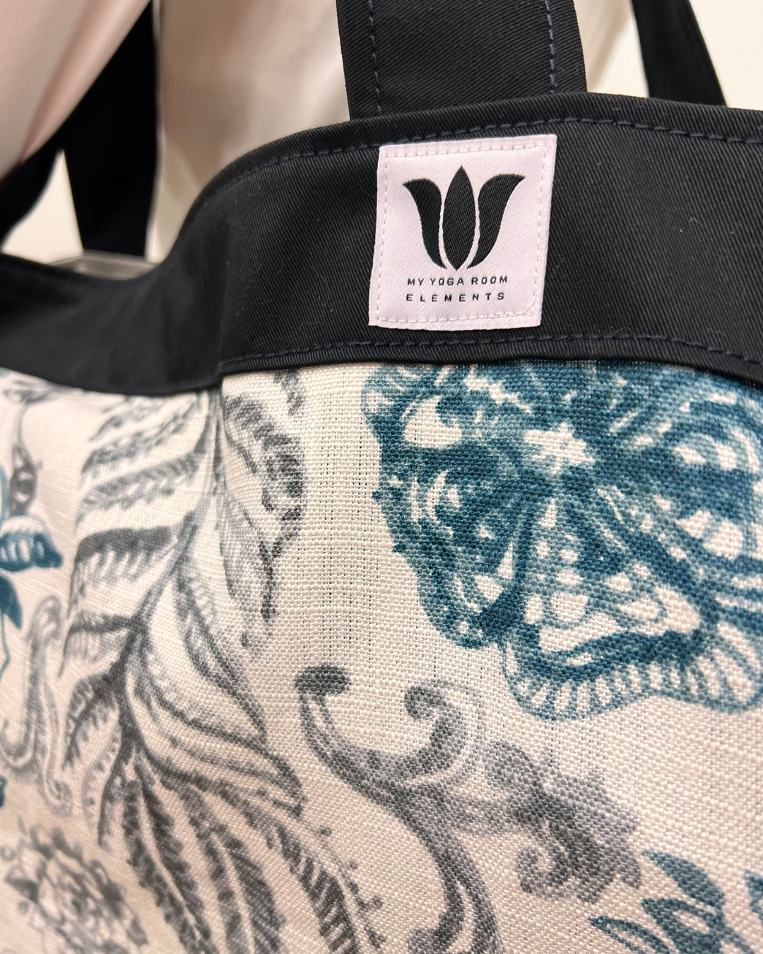 Close-up of a tote bag with a black top handle and a light beige fabric body featuring a blue-gray floral and leaf pattern. A white brand label reading 'MY YOGA ROOM ELEMENTS' is stitched onto the black trim.