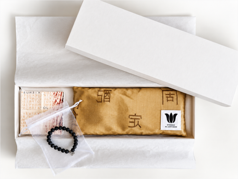 An open white gift box containing a gold fabric eye pillow with exotic characters, a small booklet, a white organza bag with a black beaded bracelet, and a white tissue. The lid rests partly open above the box.