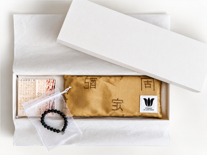 An open white gift box containing a gold fabric eye pillow with exotic characters, a small booklet, a white organza bag with a black beaded bracelet, and a white tissue. The lid rests partly open above the box.