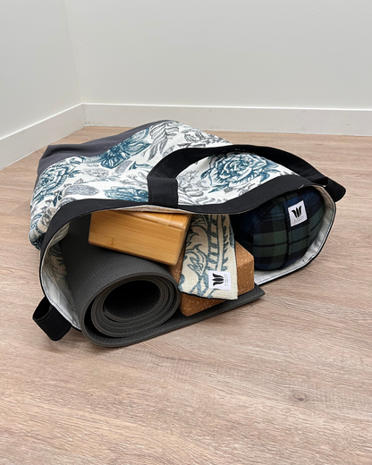 A yoga travel bag with a blue and white toile pattern, shown open on a light wood floor with a rolled yoga mat, wooden blocks, and a plaid blanket inside.